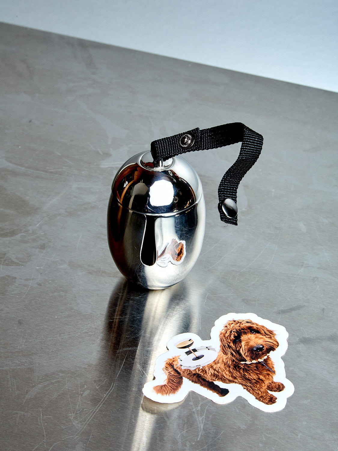 Metallic dog waste bag carrier with a black strap on a reflective surface, next to a sticker of a dog.