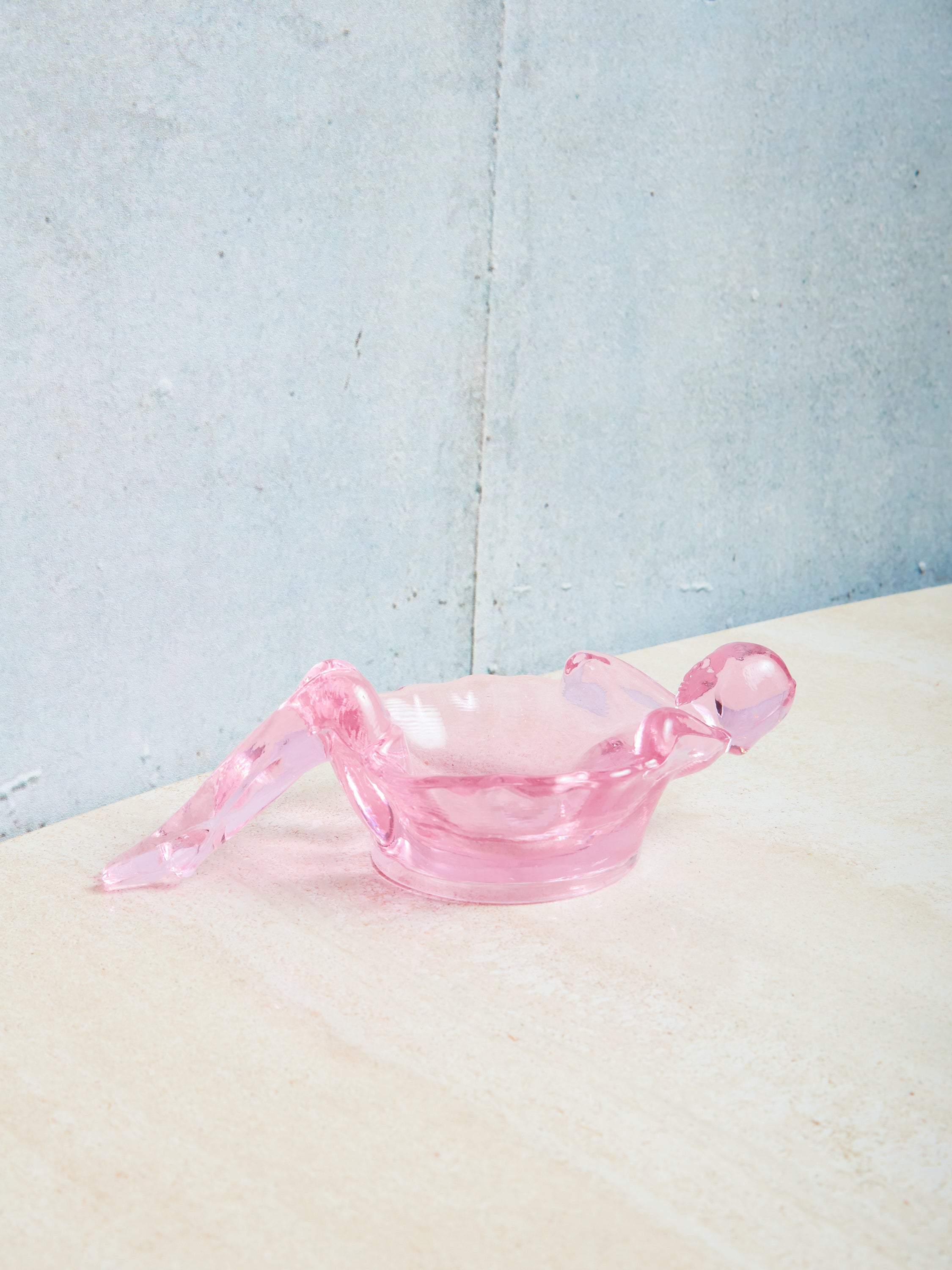 Pink glass bathing lady dish on a textured surface with a light blue background