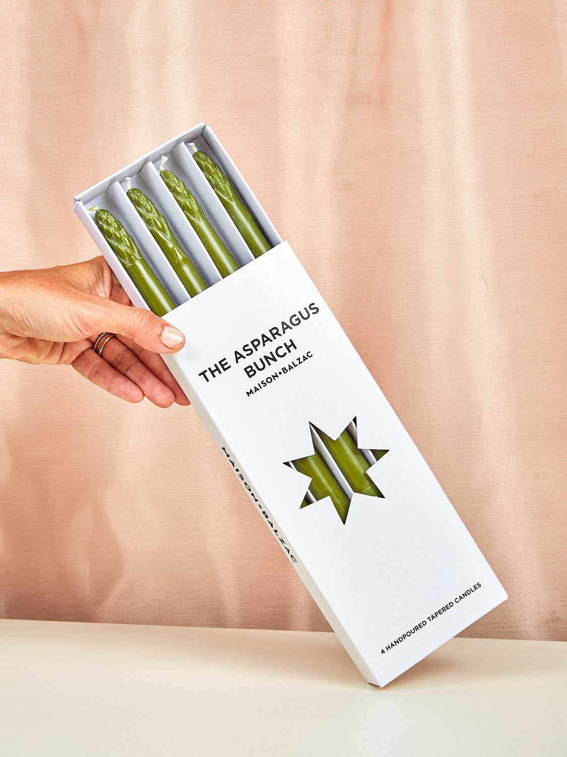 Hand holding a box of 'The Asparagus Bunch' candles against a beige background