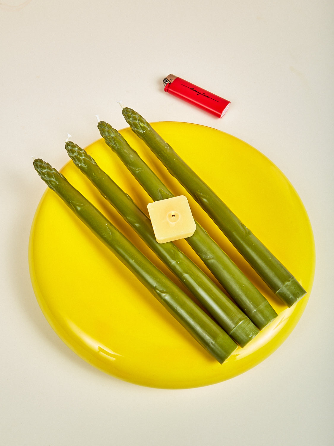Green asparagus candles on a yellow plate with a square pad of butter candle and a red lighter on a beige background