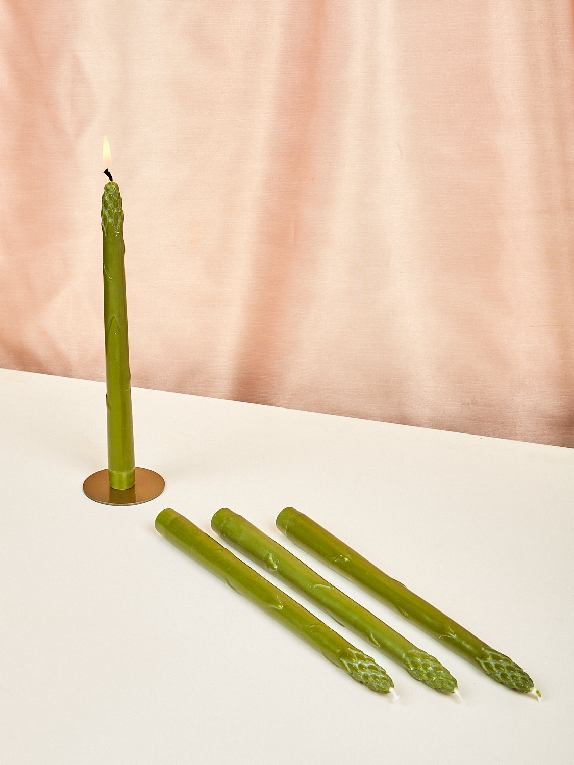 A set of green asparagus-shaped dinner candles displayed on a table with one candle lit, against a pink backdrop.