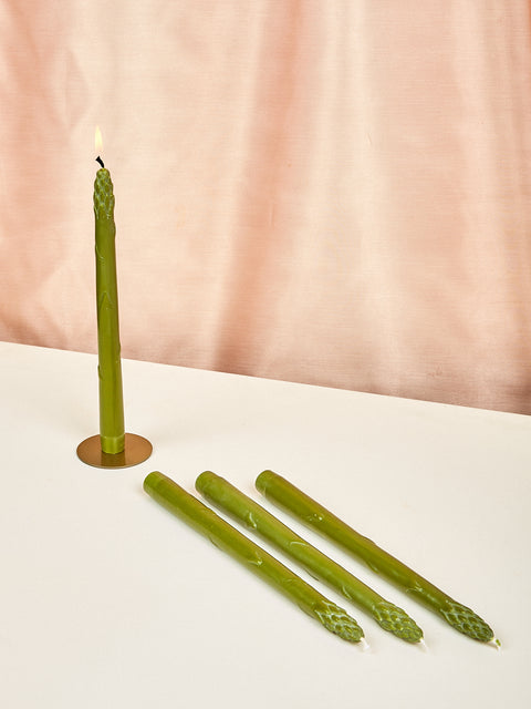 A set of green asparagus-shaped dinner candles displayed on a table with one candle lit, against a pink backdrop.