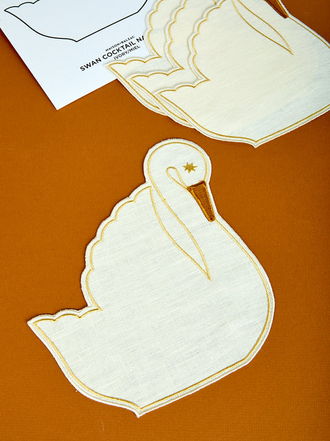 A set of white Swan-shaped cocktail napkins with a yellow outline and star detail on the head, displayed on a brown surface.