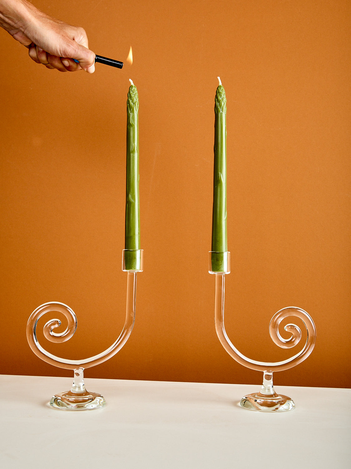 Person lighting two green candles in decorative glass candle holders against a brown background