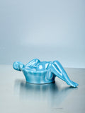 Blue glass dish of a reclining figure on a reflective surface with a light blue background