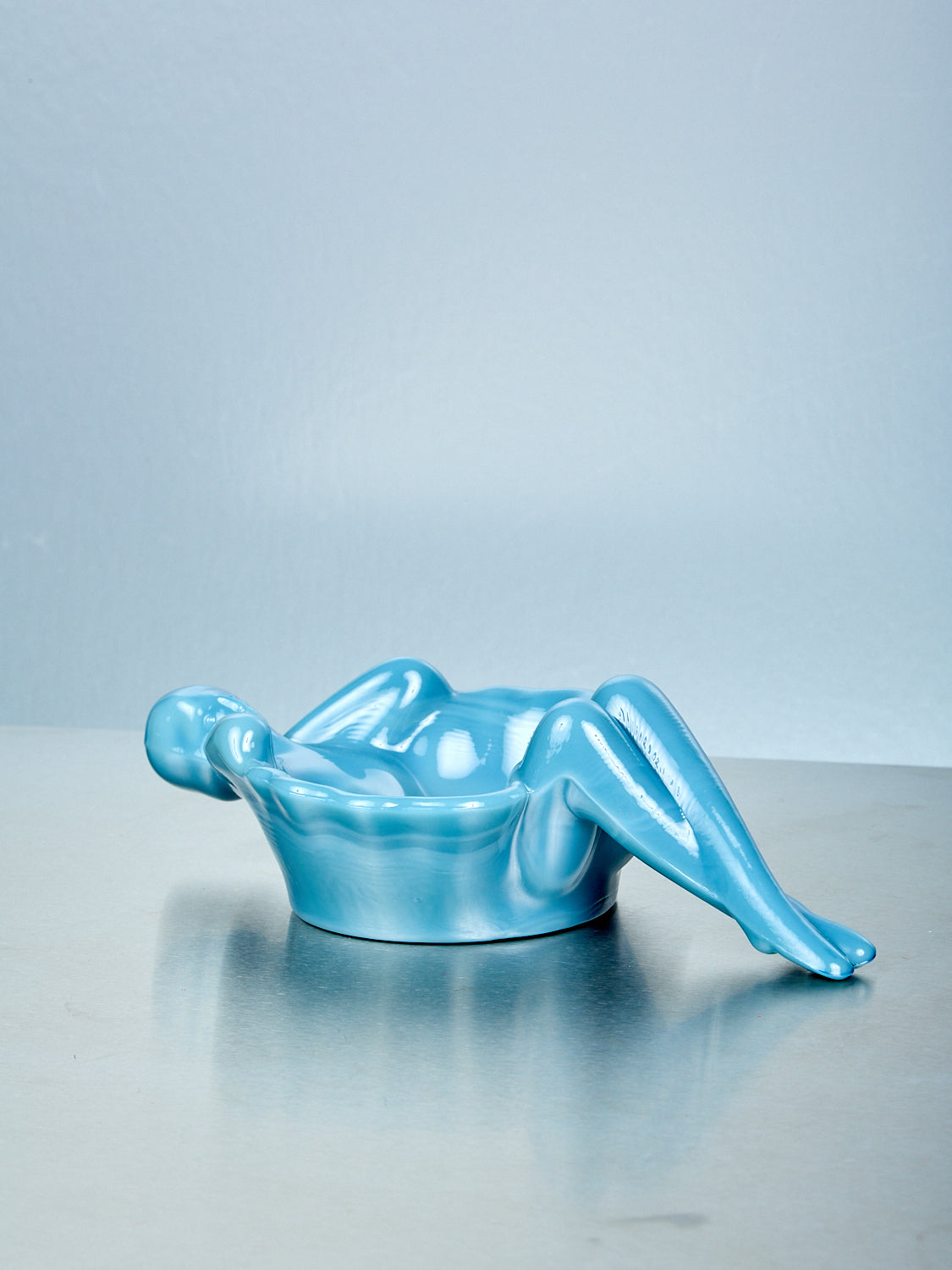 Blue glass dish of a reclining figure on a reflective surface with a light blue background