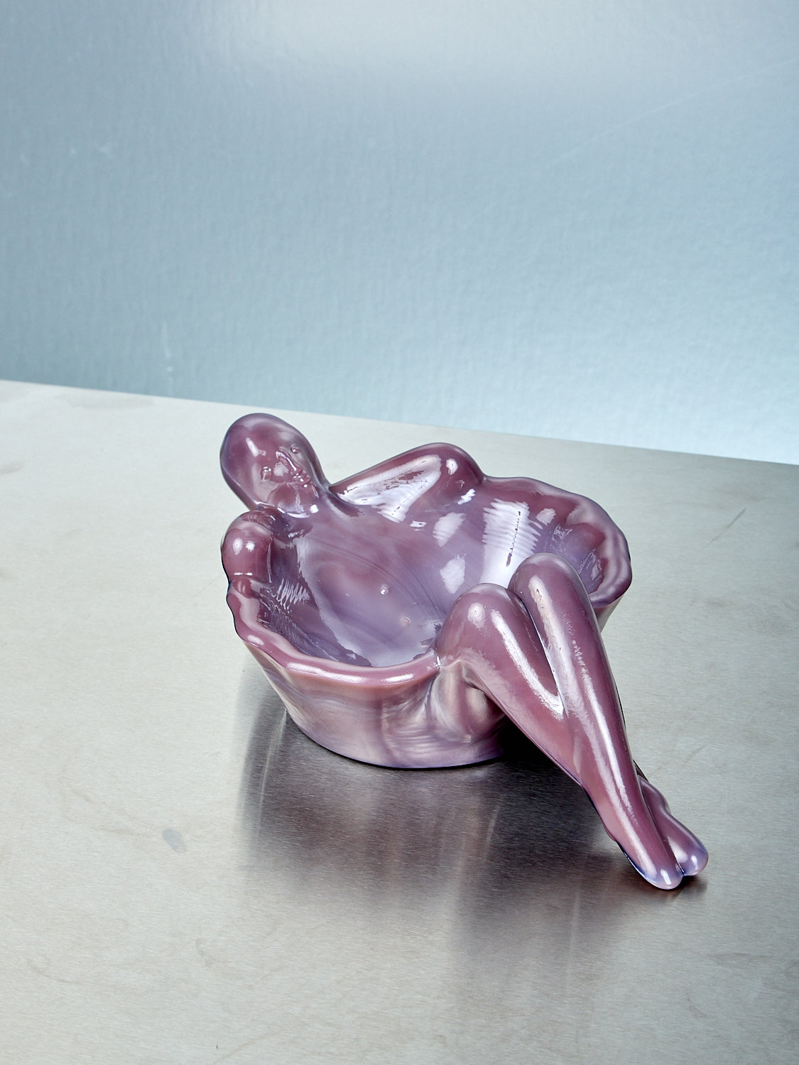 Plum purple glass sculpture of a reclining figure on a reflective surface with a light blue background