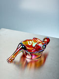 Gold multicolored glass sculpture of a reclining figure on a light gray surface with a light blue background
