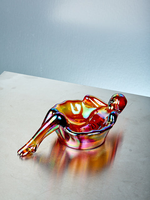 Gold multicolored glass sculpture of a reclining figure on a light gray surface with a light blue background