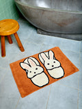 Bath mat with bunny slippers design on a bathroom floor next to a bathtub and stool.