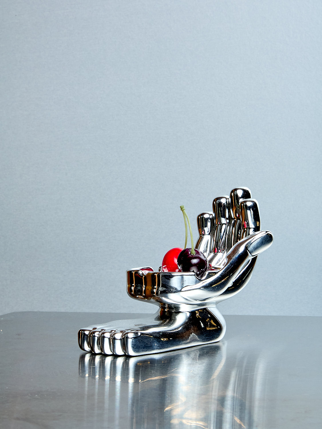 Metallic hand-shaped holder with cherries on a reflective surface against a light blue background