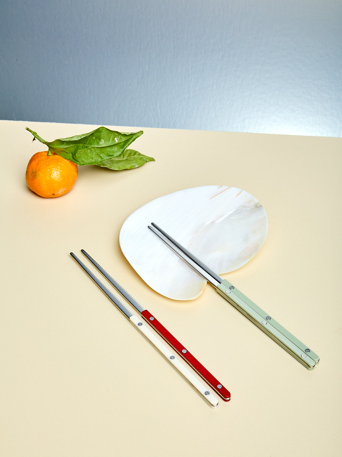 Set of three metal chopsticks on a beige surface with an orange and leaves in the background.