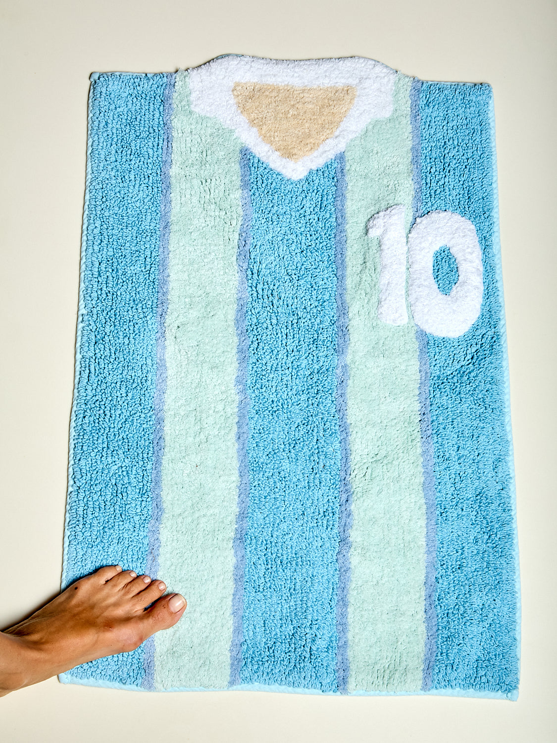 A plush bath mat designed like the Argentina national football team jersey, featuring light blue and mint green vertical stripes with a white V-neck collar and the number 10 on the chest. A human foot is visible at the bottom left for scale against a plain beige background.