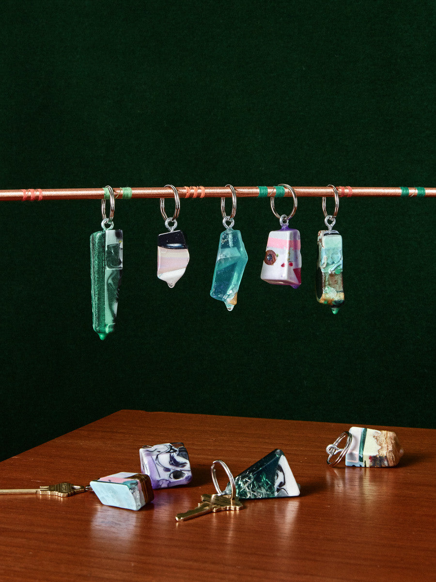 Five Nugget Keychains by Chen and Kai hang on a stick by their key ring. Below them, laying on a wooden table is 4 more keychains.